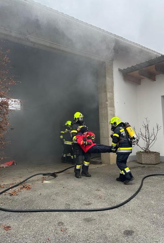 Feuerwehrleute retten eine Person aus einer brennenden Garage, mit dickem Rauch und einem Feuerlöscher sichtbar.