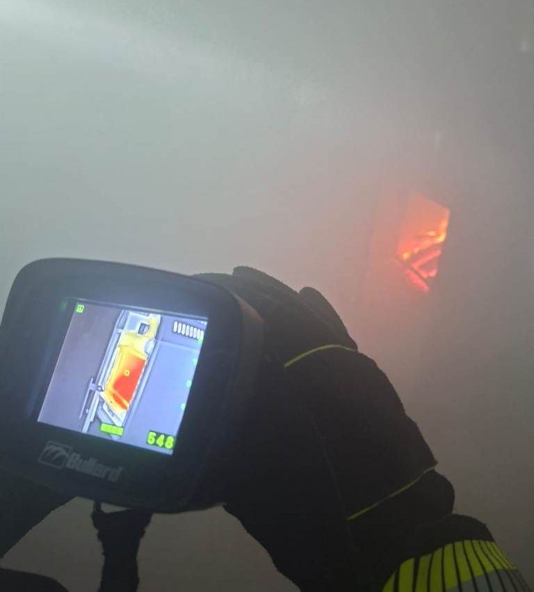 Ein Feuerwehrmann verwendet eine Bullard-Infrarotkamera, um in einer nebligen Umgebung nach Wärmequellen zu suchen und zeigt dabei eine Anzeige mit Temperaturwerten an.