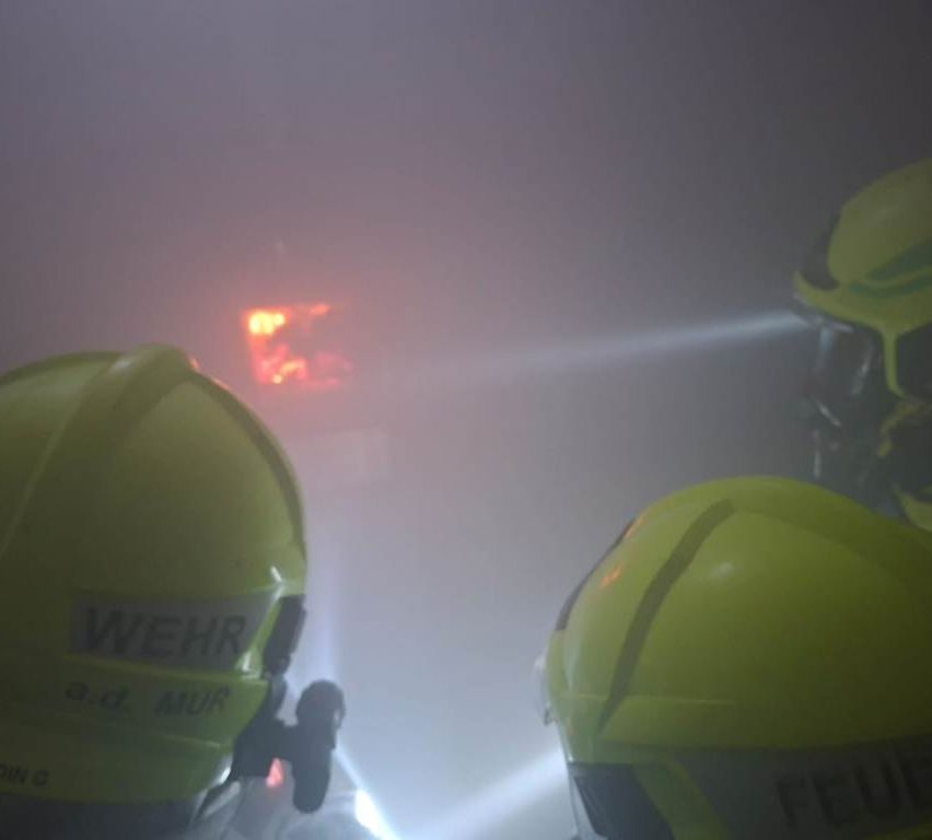 Zwei Personen in gelben Helmen mit der Aufschrift Wehr befinden sich in einem verrauchten Bereich mit einem Feuer im Hintergrund.