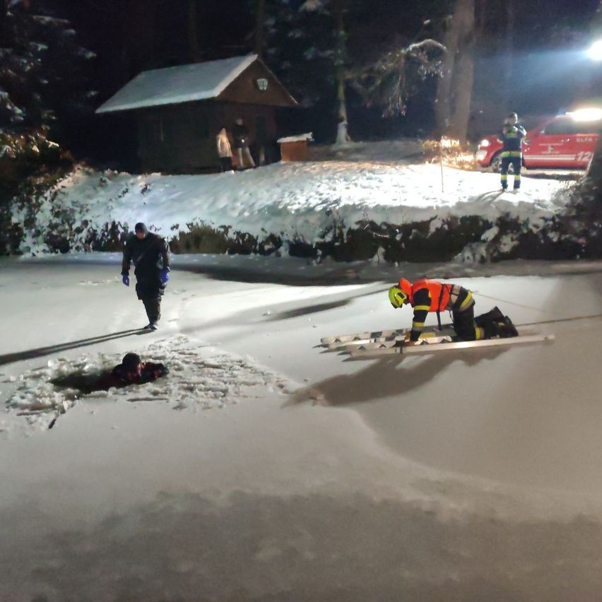 Rettungskräfte retten eine Person aus einem gefrorenen See. Eine Person ist auf Skiern, eine andere am Boden. Ein kleines Haus und ein Auto sind im Hintergrund zu sehen.