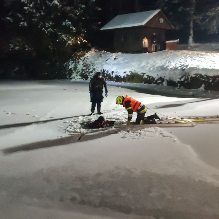 Zwei Personen stehen bei Nacht auf einem zugefrorenen See. Eine Person kniet in einer reflektierenden Weste und einem Helm, während eine andere Person liegt. Ein Haus ist im Hintergrund zu sehen.