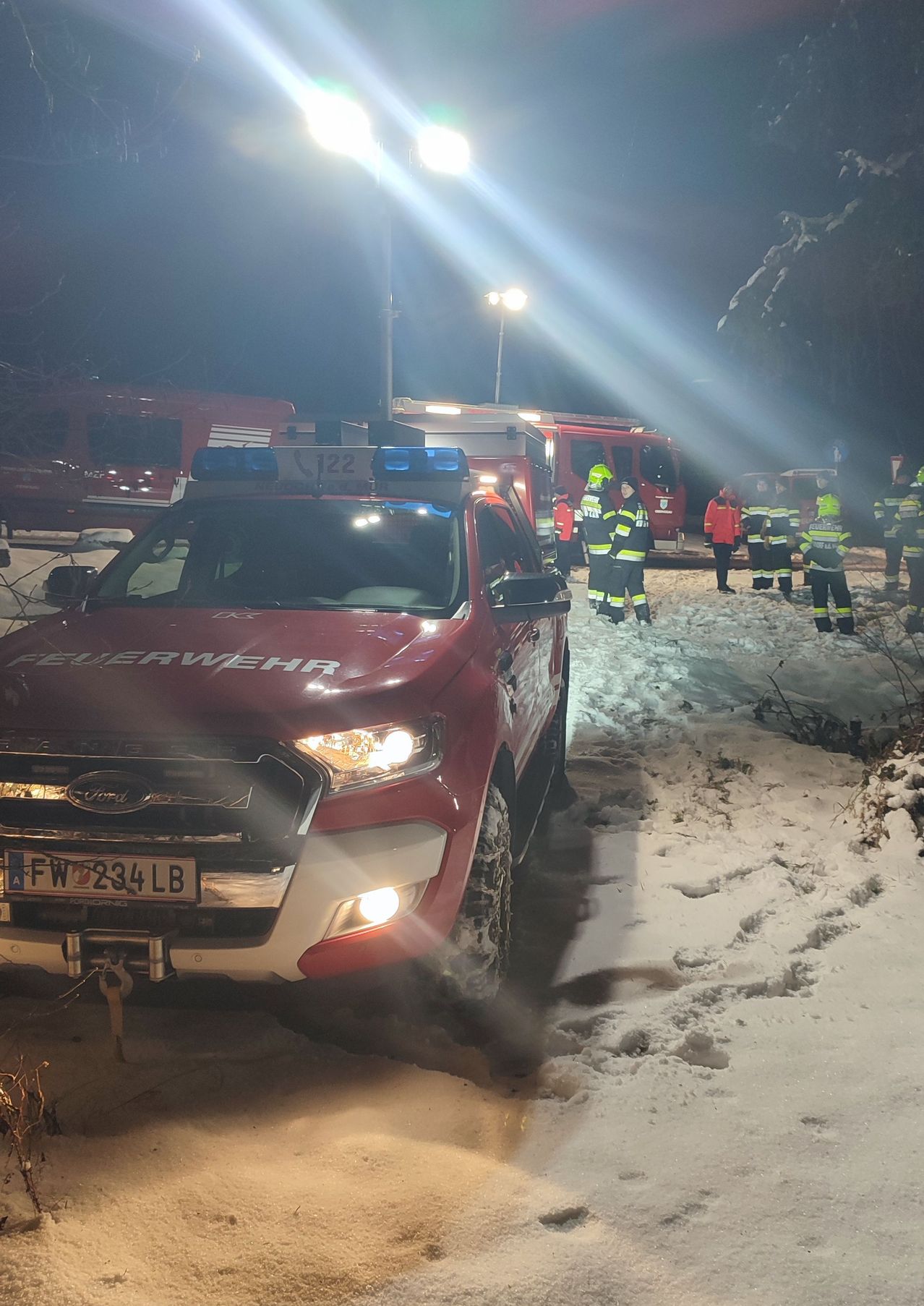Ein roter Feuerwehrwagen mit dem Kennzeichen FWZ234LB steht im Schnee. Notfallpersonal ist in der Nähe, und Feuerwehrwagen sind im Hintergrund.