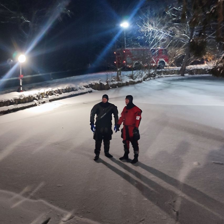 Zwei Personen stehen nachts auf einer verschneiten Straße, beleuchtet von Straßenlaternen. Ein Feuerwehrauto ist in der Nähe geparkt.