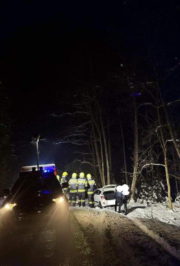 Eine Gruppe von Feuerwehrleuten mit gelben Helmen steht neben einem weißen Auto mit offenem Kofferraum auf einer verschneiten Straße bei Nacht.