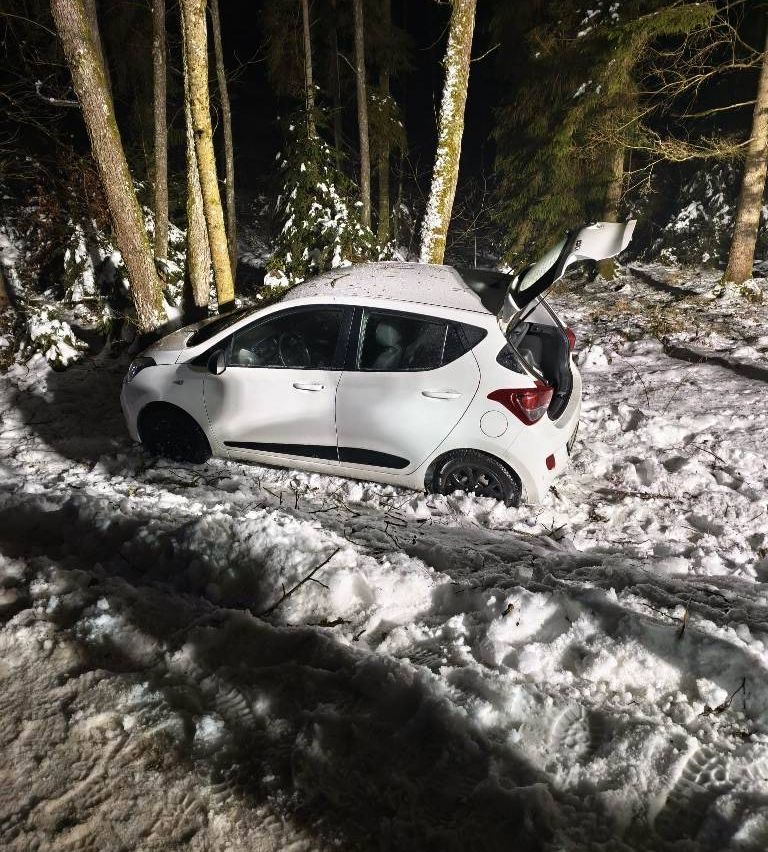 Ein weißer Wagen steckt im Schnee fest, der Kofferraum ist geöffnet, umgeben von Bäumen in einem verschneiten Bereich.