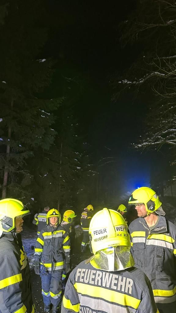 Eine Gruppe Feuerwehrleute, die gelbe Helme tragen, sind in einem verschneiten Wald bei Nacht versammelt. Sie reagieren wahrscheinlich auf einen Notfall.