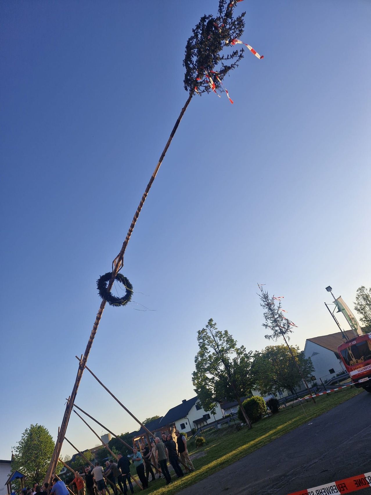 A tall wooden pole with a wreath at the top and some red ribbons, near a red fire truck and houses under a clear blue sky.