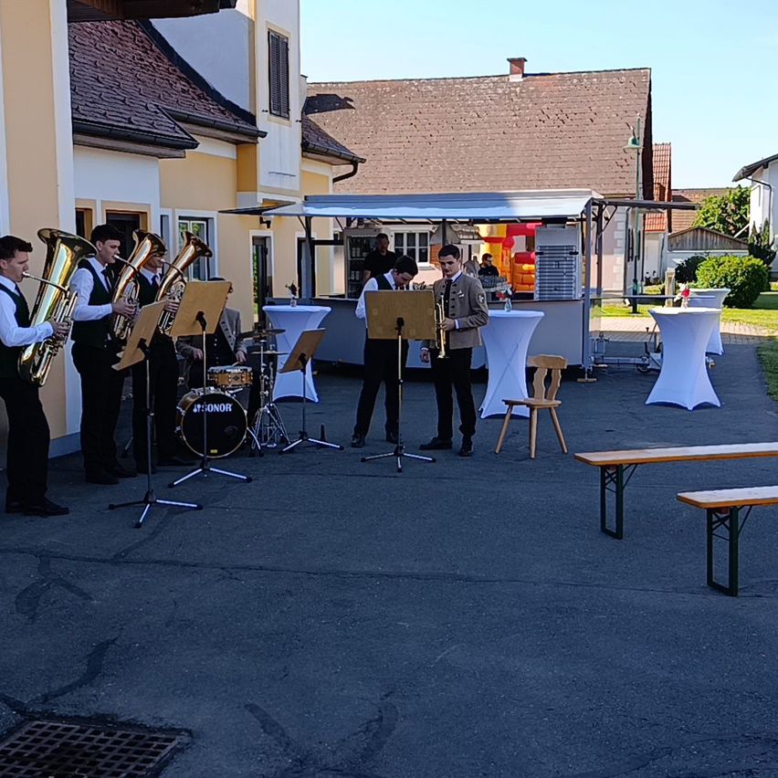 A group of musicians plays brass instruments in an outdoor setting. A food truck is nearby, with tables and chairs set up. The setting is outdoors with buildings in the background.