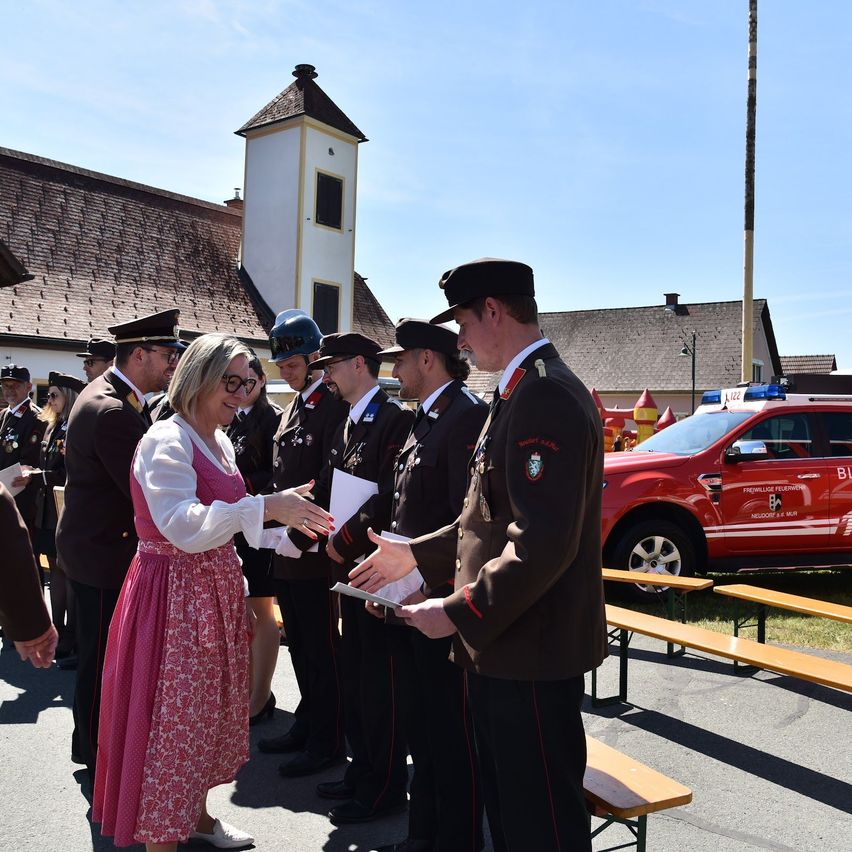 A woman in a traditional dress shakes hands with a group of men in uniform, standing in front of a church and a red vehicle.