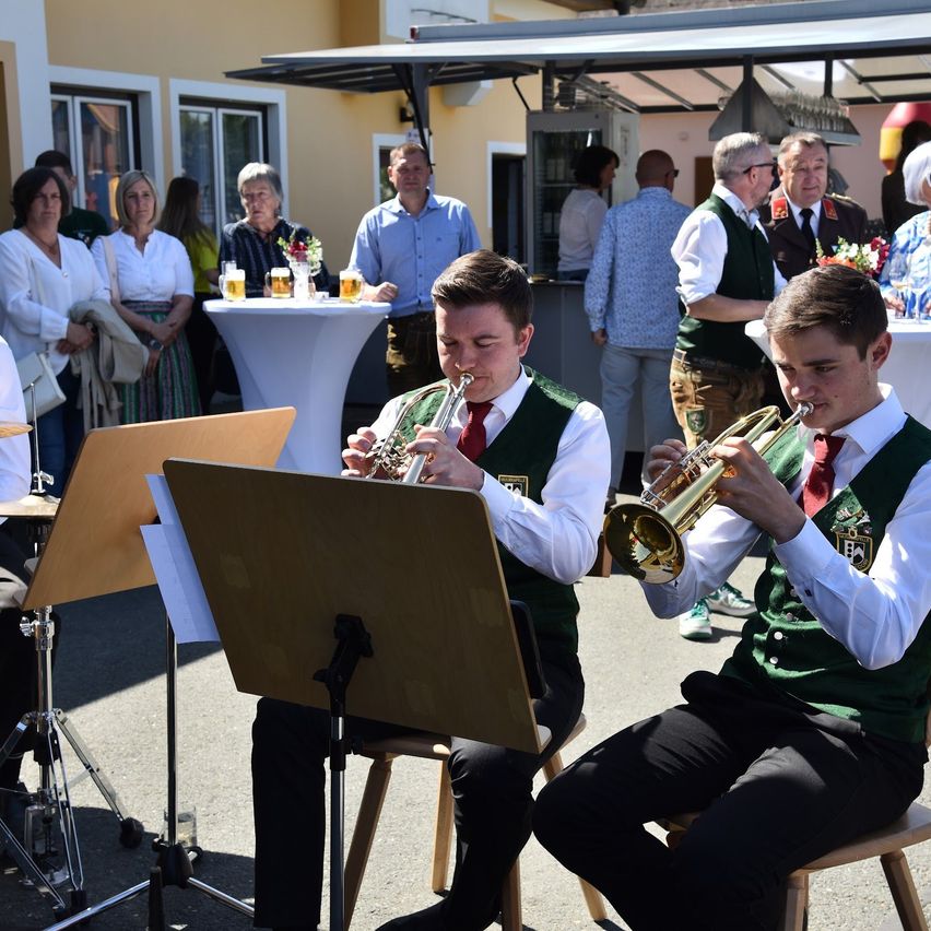 Two young men play trumpets outdoors in front of a crowd. Behind them, a group of people are standing around a table with drinks.