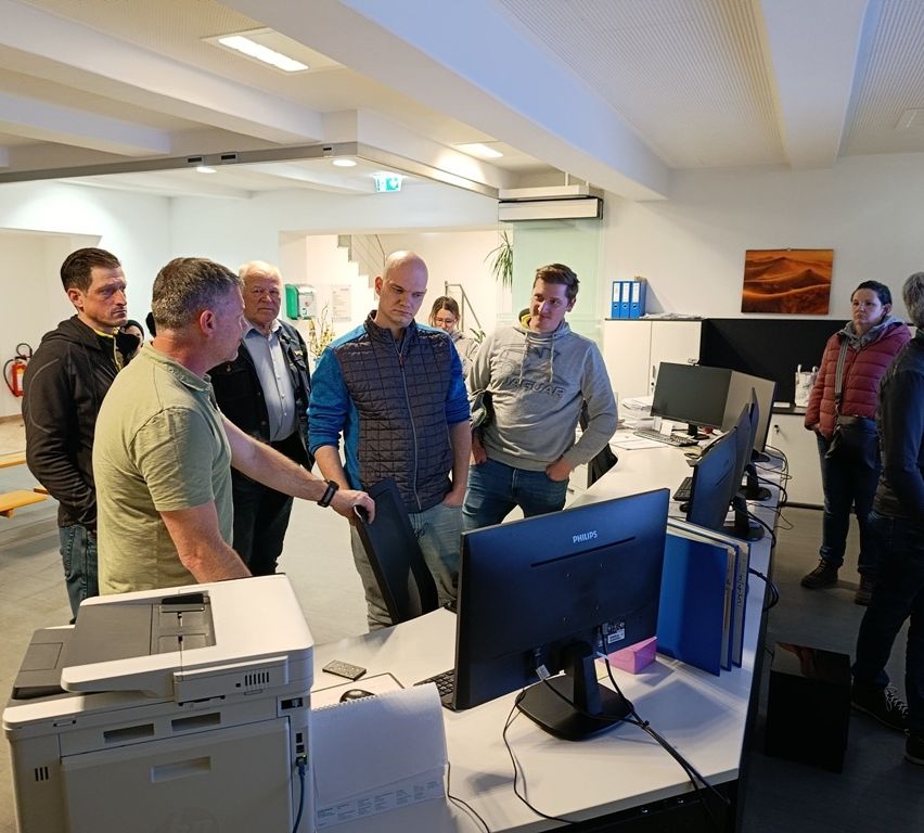 Eine Gruppe von Männern versammelt sich um einen Bürocomputer, möglicherweise um arbeitsbezogene Angelegenheiten zu besprechen. Ein Drucker und ein Monitor sind auf dem Schreibtisch zu sehen.