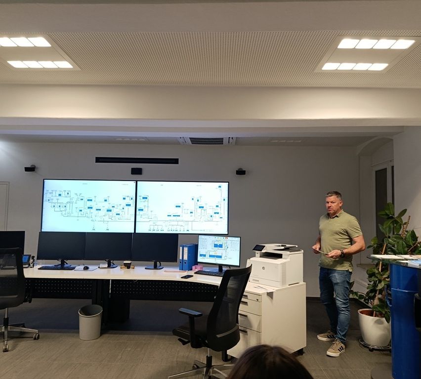 Ein Mann steht in einem Büroraum mit drei großen Monitoren und hält eine Präsentation. Der Raum hat weiße Wände und einen Holzboden.