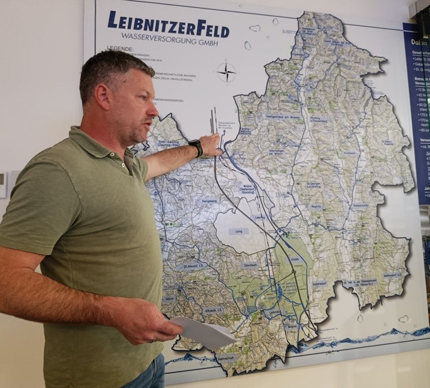 Ein Mann steht vor einer großen Karte und zeigt mit der linken Hand auf einen bestimmten Bereich, während er ein Papier in der rechten Hand hält. Die Karte hat einen blauen Rand und ist mit "LEIBNITZERFELD WASSERVERSORGUNG GMBH" beschriftet.