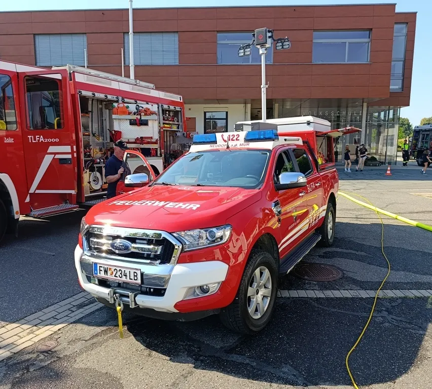 Ein roter Ford Ranger Pickup-Truck mit Notfallleuchten auf dem Dach ist vor einem Gebäude geparkt. Ein Feuerwehrmann steht neben einem Feuerwehrwagen.