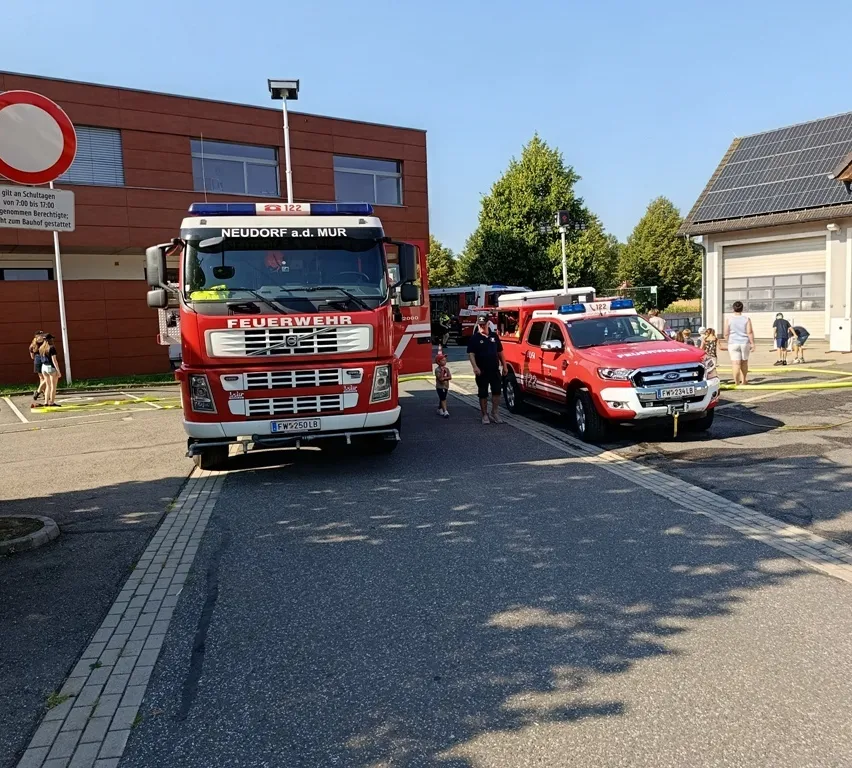 Zwei Feuerwehrfahrzeuge sind am Straßenrand geparkt, neben einem steht ein Feuerwehrmann. Hinter ihnen befindet sich ein Gebäude mit Solarmodulen, und mehrere Menschen gehen herum.