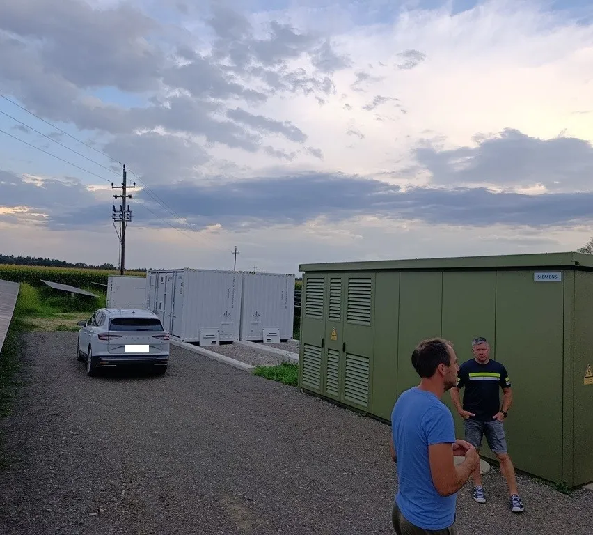 Zwei Männer stehen vor einem grünen Siemens-Container, mit einem geparkten Auto und weißen Containern im Hintergrund.