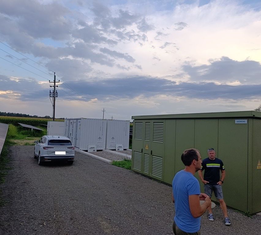 Two men are standing in front of a green Siemens container, with a parked car and white containers in the background.