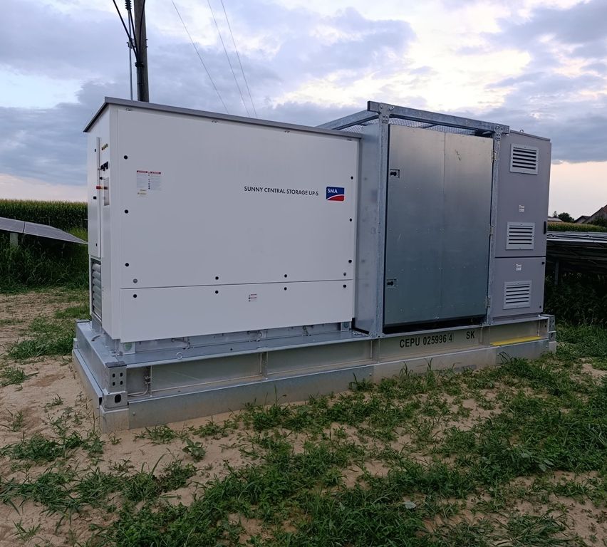 A large white Sunny Central Storage UPS system is installed on a concrete base in an open field with green grass and a cloudy sky.