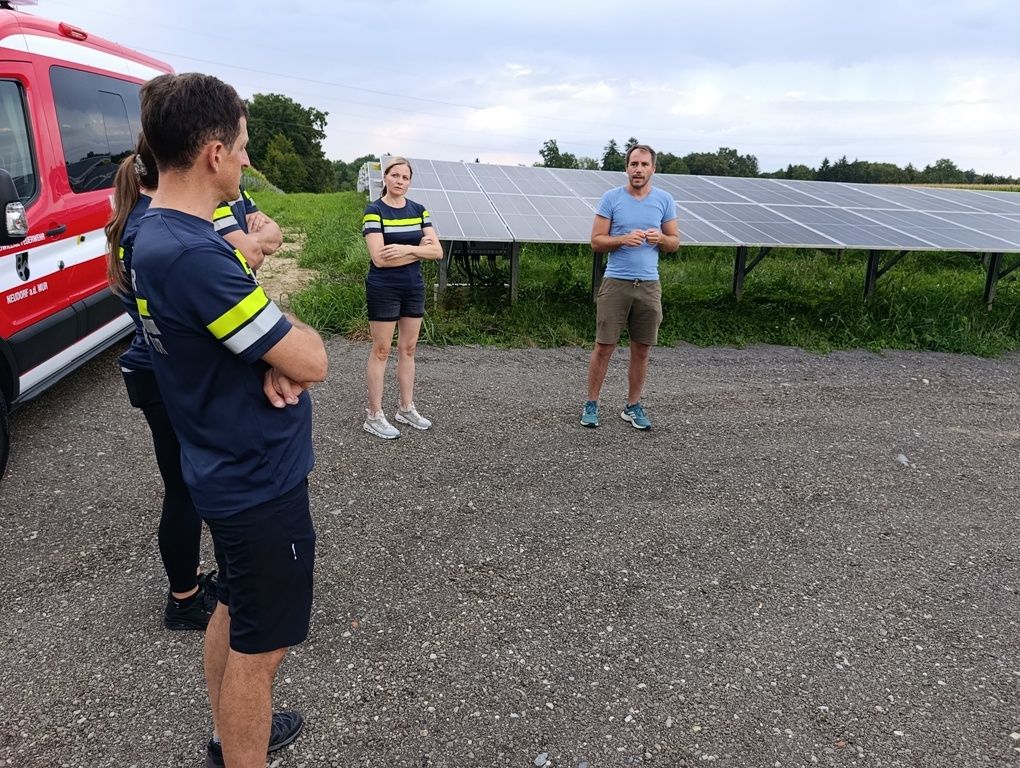 A group of people dressed in blue uniforms are standing in front of a solar panel installation. The man in the center is speaking to the group.