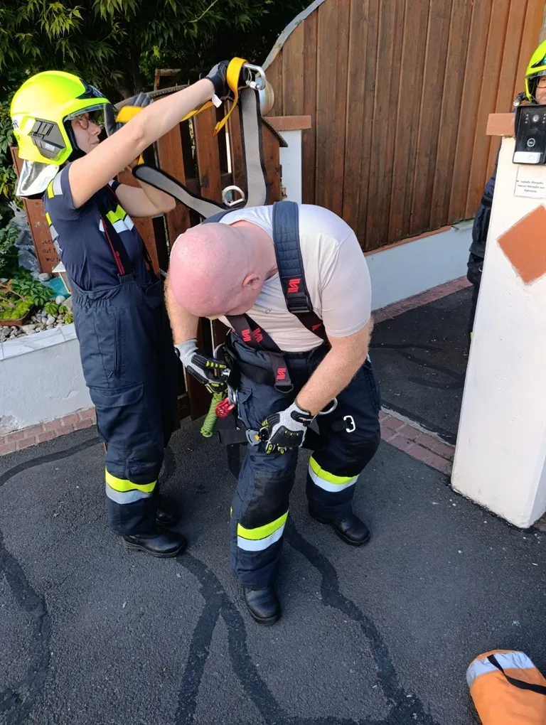 Ein Feuerwehrmann justiert den Gurt eines anderen Feuerwehrmanns vor einem Holzhaus.