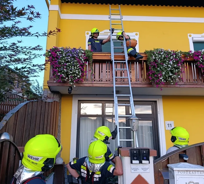 Feuerwehrleute in gelben Helmen und Ausrüstung klettern auf einer Leiter auf einem Balkon mit Blumenkästen. Ein Feuerwehrmann hält eine Kürbis.