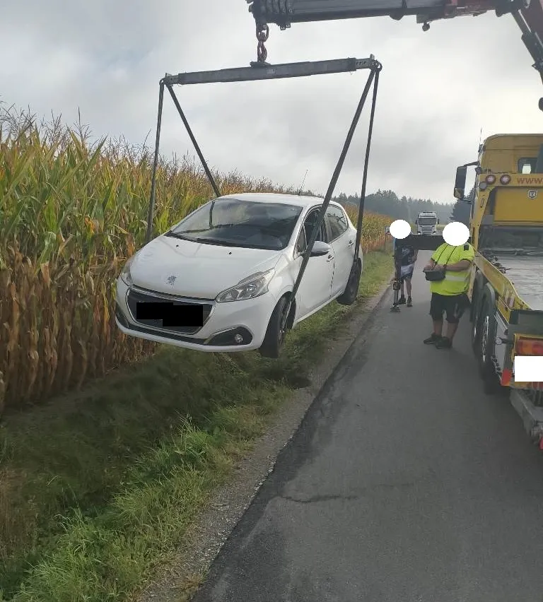 Ein weißer Wagen wird von einem Abschleppwagen am Straßenrand angehoben, in der Nähe eines Maisfeldes. Zwei Personen stehen in der Nähe, eine trägt eine reflektierende Weste.
