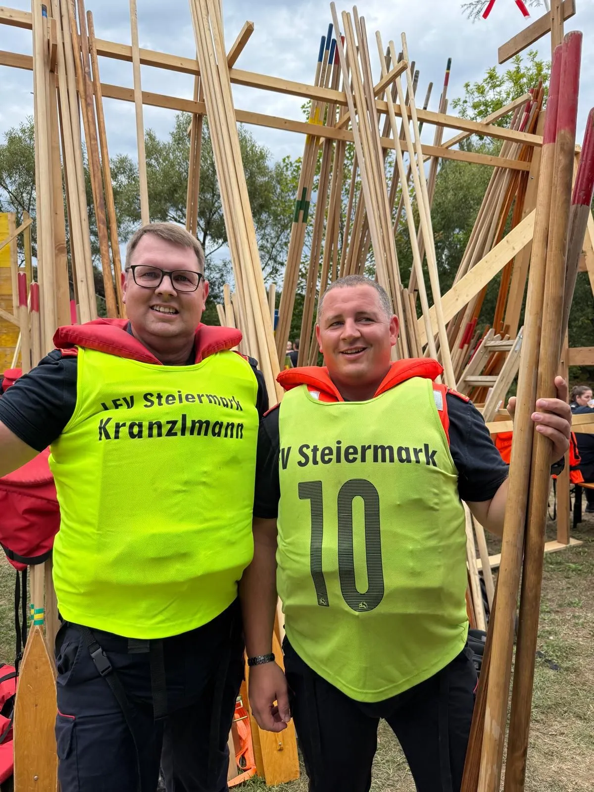 Zwei Männer in Sicherheitswesten, einer mit Kranzmann und der andere mit Nummer 10, stehen vor einer Holzstruktur.