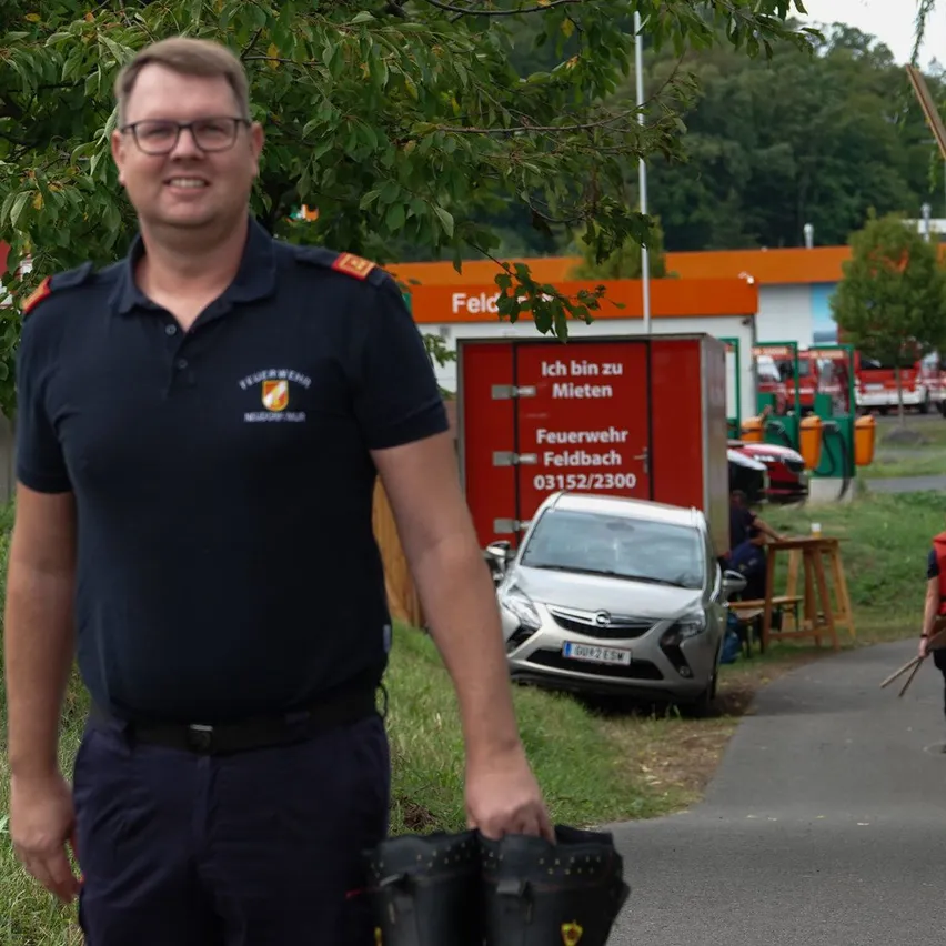 Ein Feuerwehrmann in Uniform steht vor einem Feuerwehrhaus und hält ein Paar Stiefel. Dahinter ist ein Feuerwehrwagen geparkt, und eine Person sitzt an einem Tisch.