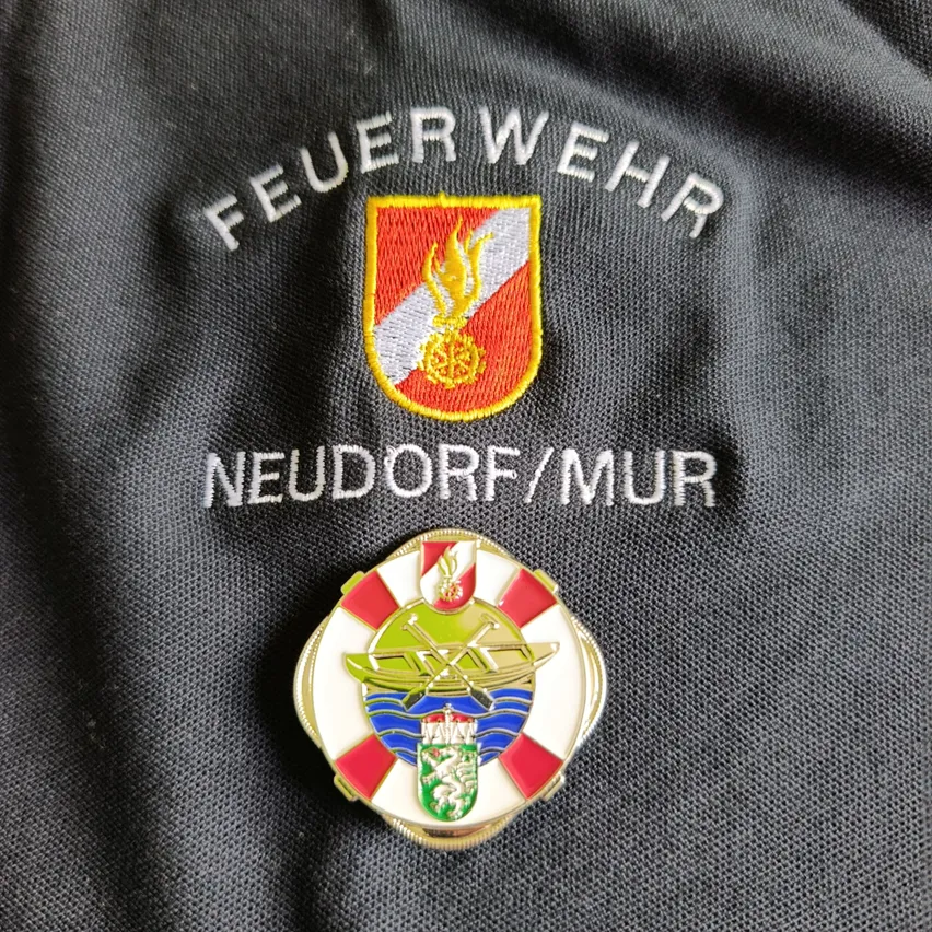 Ein Nahaufnahme eines Feuerwehrmann-Uniforms mit den Worten 'FEUERWEHR' und 'NEUDORF/MUR' aufgestickt. Zwei Abzeichen sind an der Uniform befestigt, eines mit einem Feuersymbol und das andere mit einem Schilddesign.