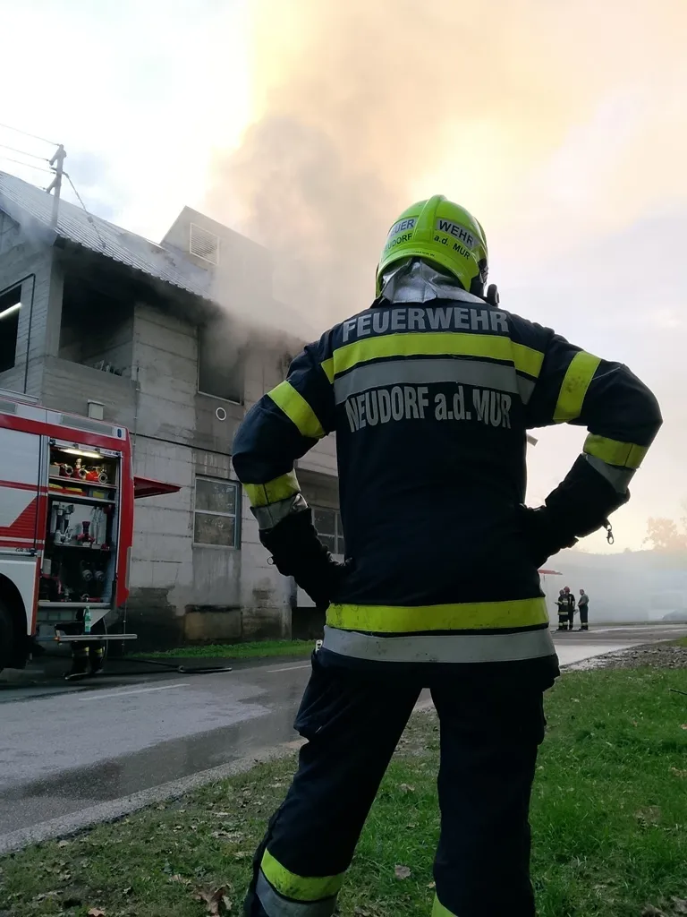 Ein Feuerwehrmann in voller Ausrüstung steht mit dem Rücken zur Kamera und sieht einem brennenden Gebäude mit aufsteigendem Rauch zu.