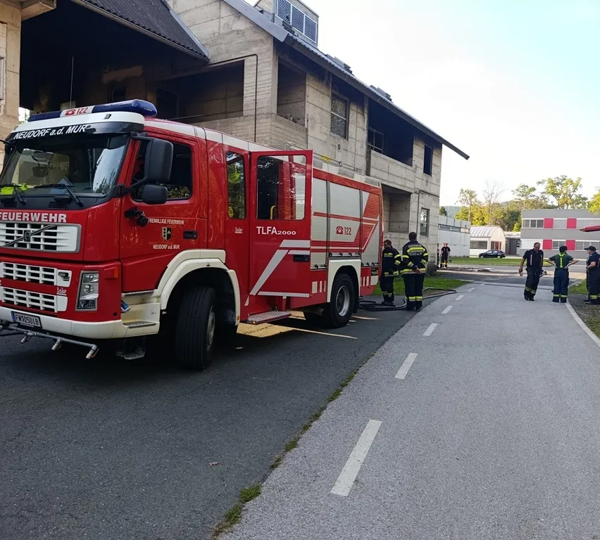 Ein roter Feuerwehrwagen ist vor einem Gebäude geparkt, in der Nähe befinden sich Feuerwehrleute.