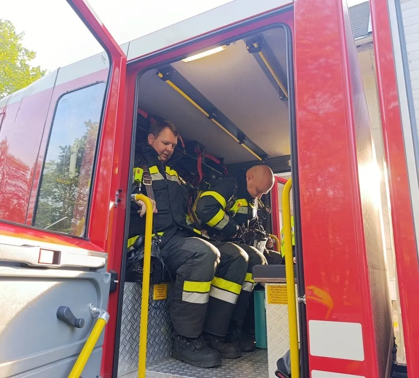 Zwei Feuerwehrleute sitzen in einem roten Feuerwehrauto mit offener Tür, einer lächelt und der andere schaut nach unten.