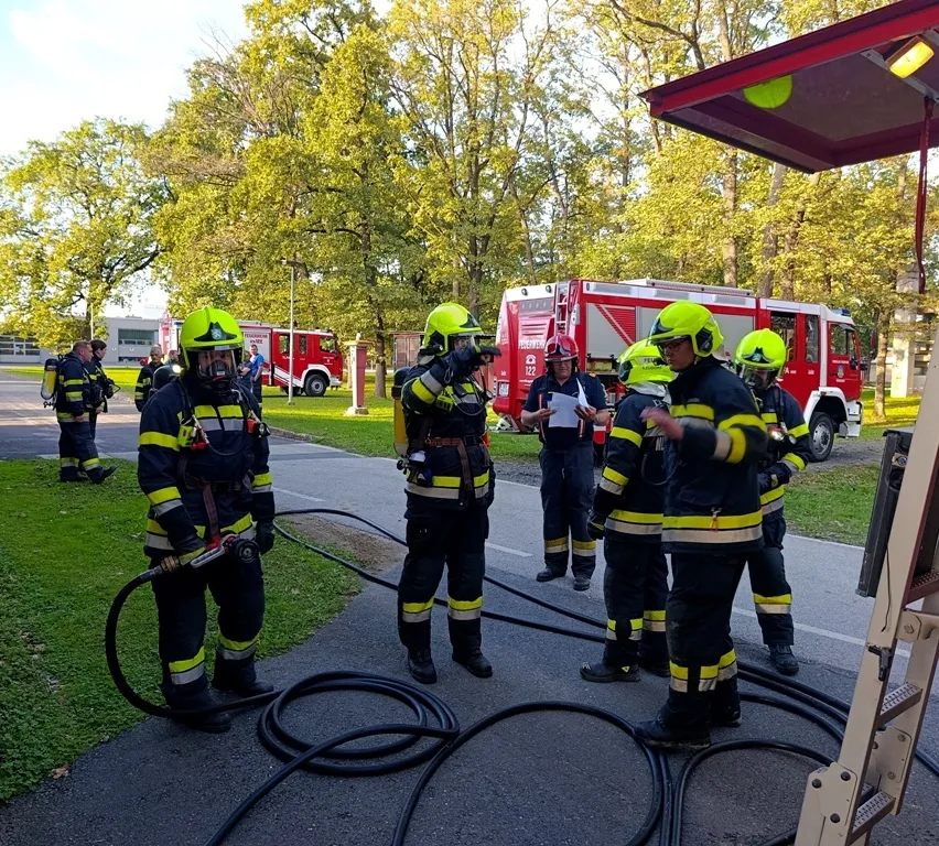 Eine Gruppe von Feuerwehrleuten in voller Ausrüstung versammelt sich um einen Feuerwehrwagen, einer hält eine Feuerwehrschläuche und ein anderer liest ein Dokument.