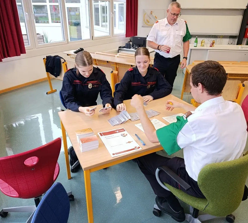 Zwei Frauen in Uniform sitzen an einem Tisch und studieren Karten, während ein Mann in einem weißen Hemd zusieht. Ein Mann in einem grünen Hemd steht hinter ihnen und hält ein Getränk.