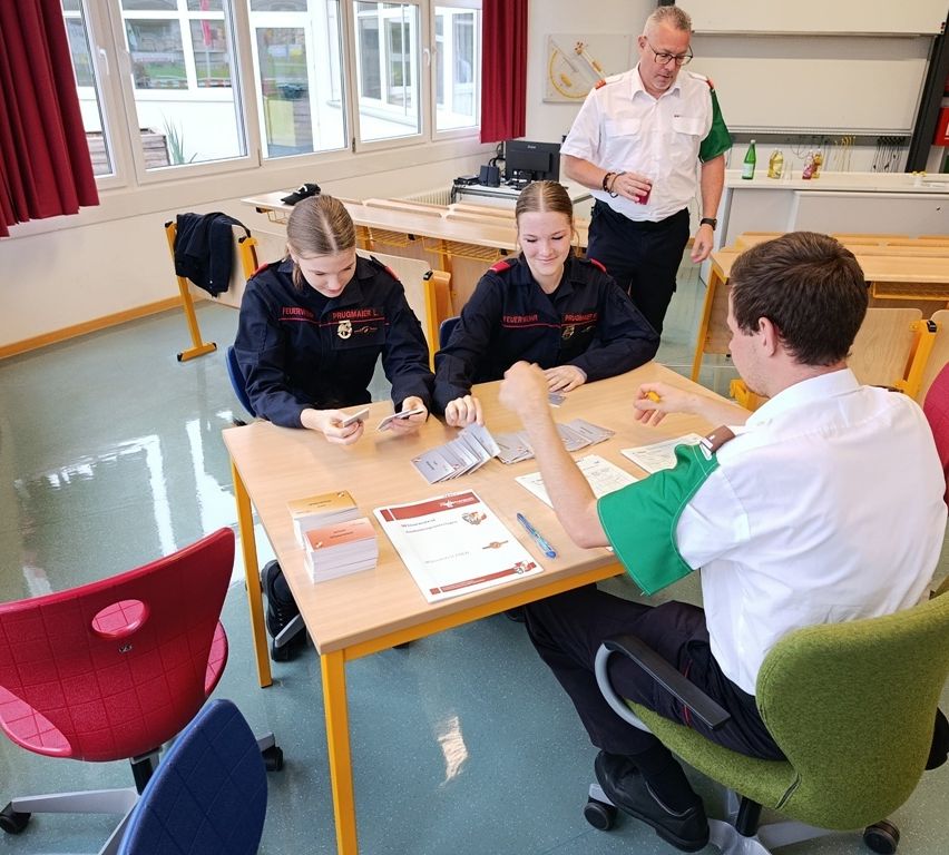 Zwei Frauen in Uniform sitzen an einem Tisch und studieren Karten, während ein Mann in einem weißen Hemd zusieht. Ein Mann in einem grünen Hemd steht hinter ihnen und hält ein Getränk.