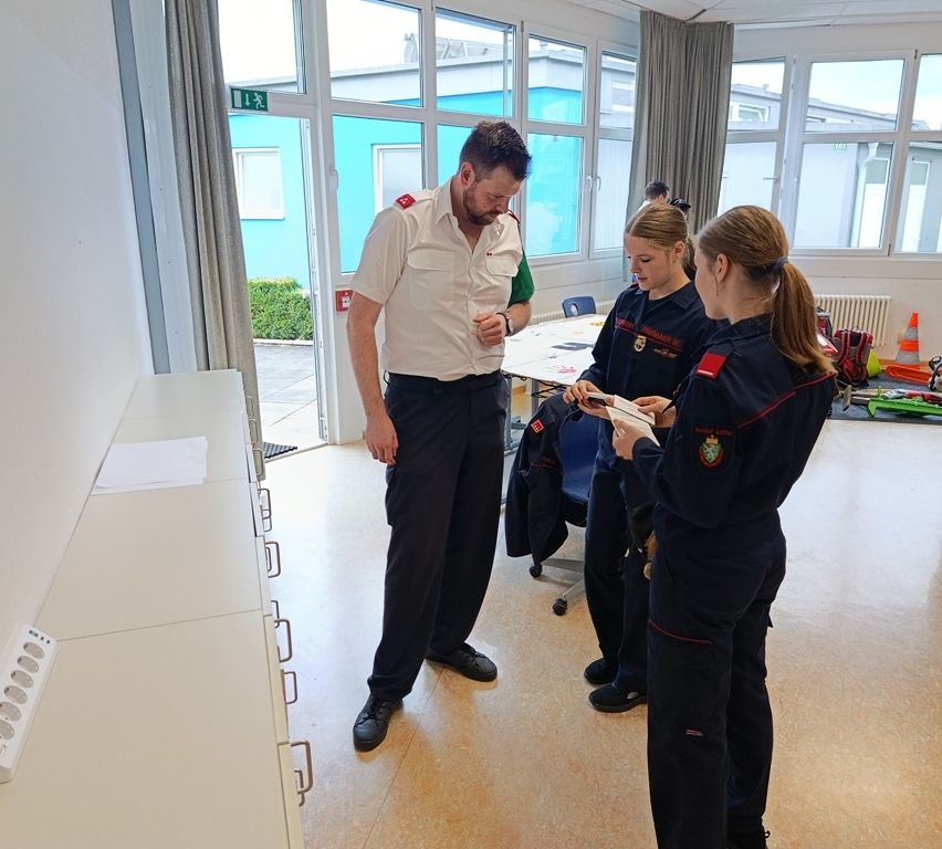 Ein Mann in weißer Uniform steht in einem Raum mit drei Frauen in blauen Uniformen, eine hält ein Buch. Sie sind in der Nähe eines Tisches mit Papieren und einer Tür.