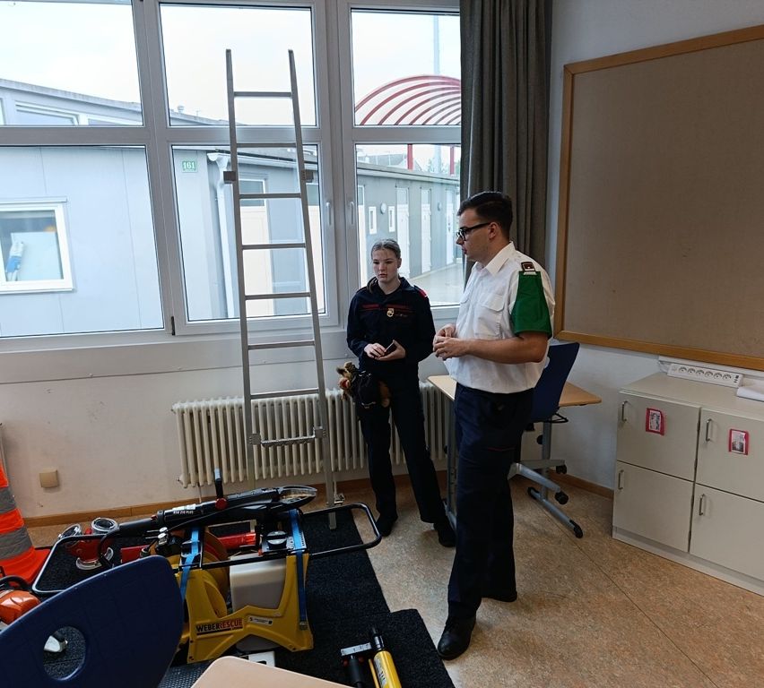 Ein Mann in Uniform steht in einem Raum mit einer Frau in Uniform, beide betrachten ein Gerät. Eine Leiter und ein Heizkörper sind im Hintergrund.