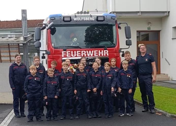 Eine Gruppe von Kindern in Feuerwehruniformen posiert vor einem roten Feuerwehrauto mit der Aufschrift 'Feuerwehr'. Sie stehen vor einem Gebäude, neben ihnen steht ein Mann in Uniform.