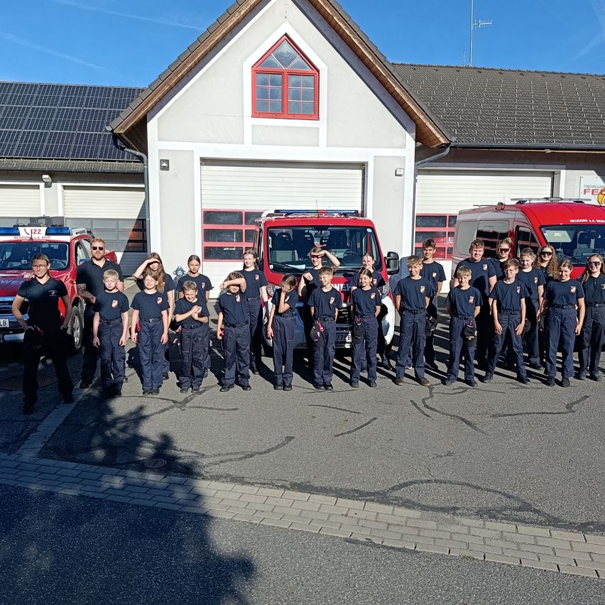 Eine Gruppe von Kindern in blauen Uniformen steht vor einer Feuerwache. Einige Erwachsene sind dahinter, und zwei rote Fahrzeuge sind geparkt. Das Gebäude hat Solarmodule und rote Fenster.