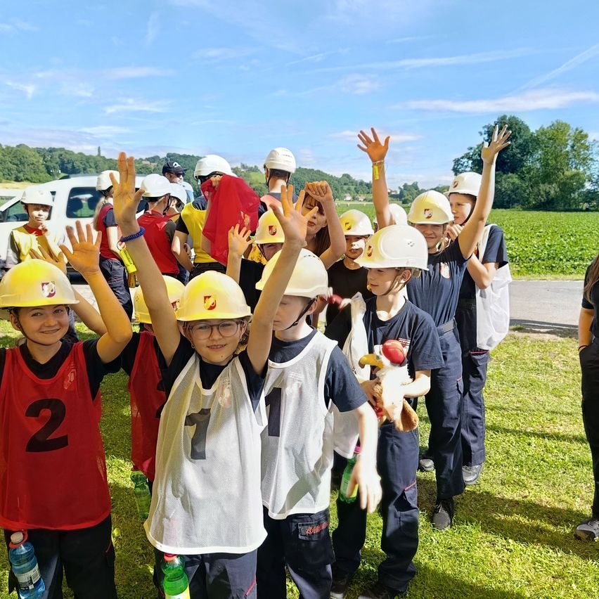 Gruppe von Kindern in gelben Schutzhelmen und nummerierten Trikots heben ihre Hände in einem Feld. Hinter ihnen steht ein weißer Van und grüne Bäume.