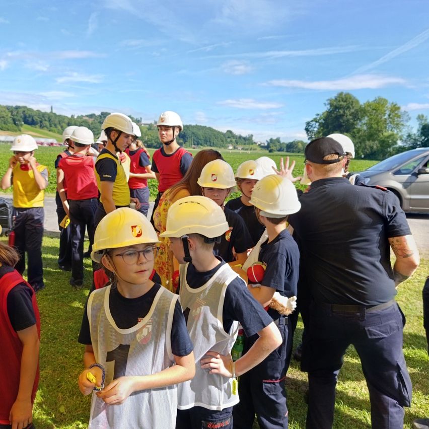 Eine Gruppe von Kindern und Erwachsenen in Sicherheitswesten und Helmen steht auf Gras, möglicherweise für eine Sicherheitsschulung. Ein Erwachsener in Uniform spricht mit den Kindern.