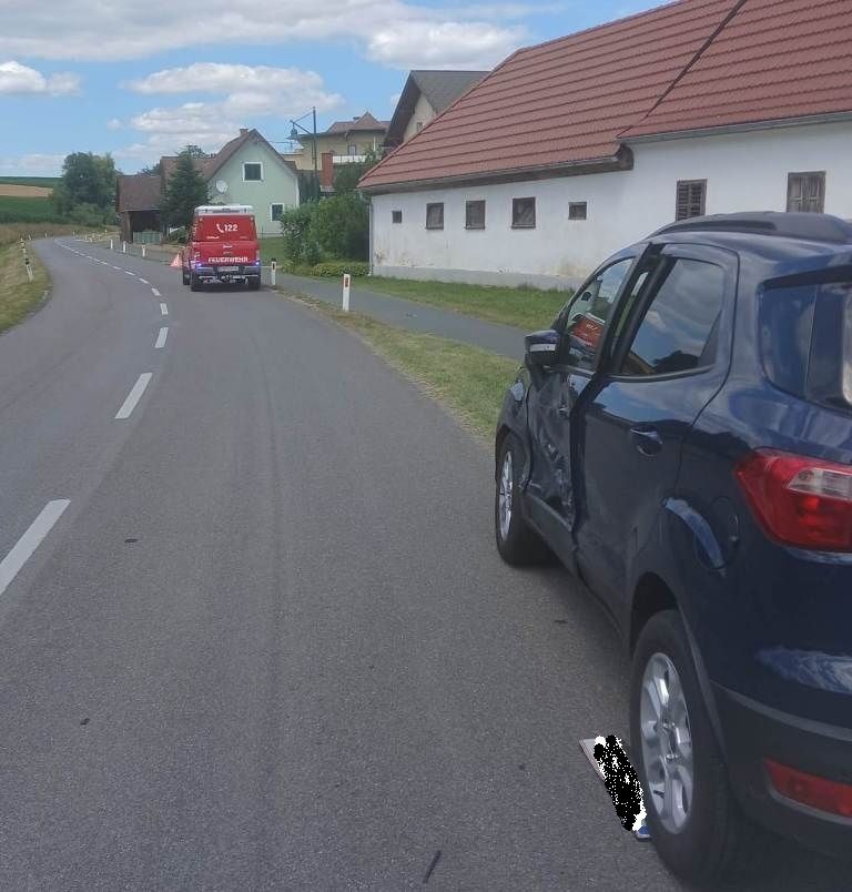 A red van with a roof rack is driving down a rural road. A blue car is parked on the side of the road, with its door open.