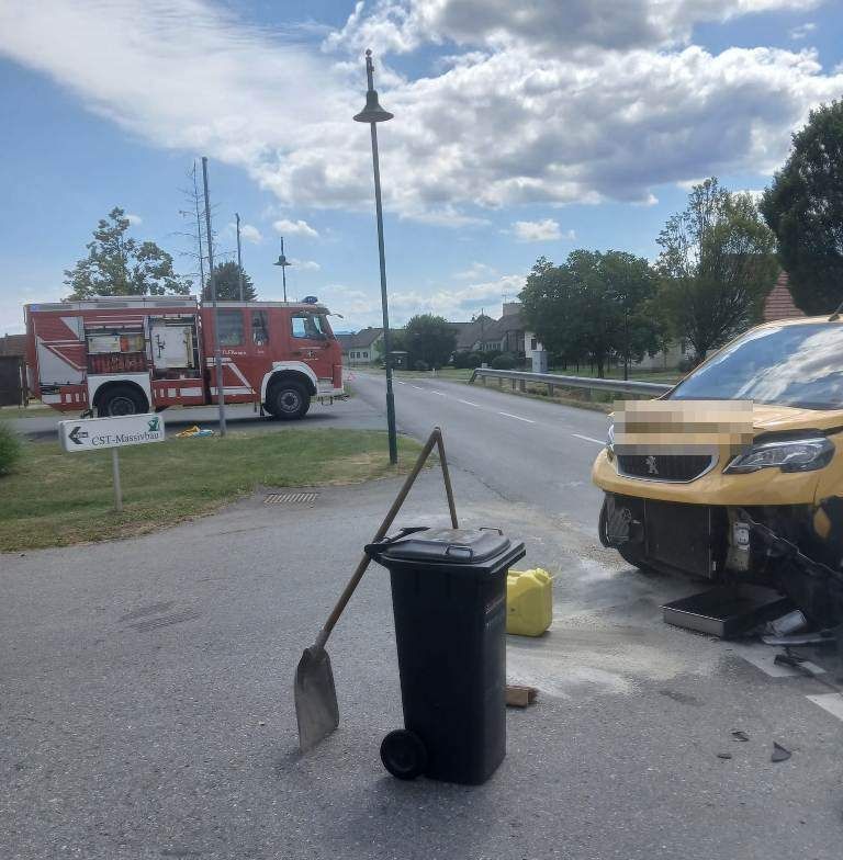 A yellow car has crashed into a trash can on the road, with a fire truck and a shovel nearby. The car's front is damaged, and the surroundings include trees and houses.