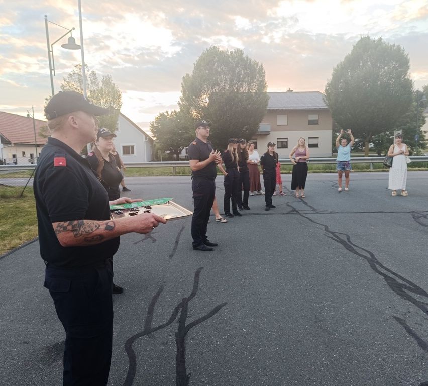 A group of uniformed individuals, possibly police officers, gather in a parking lot. A man in front holds a board with items. Trees and buildings are in the background.