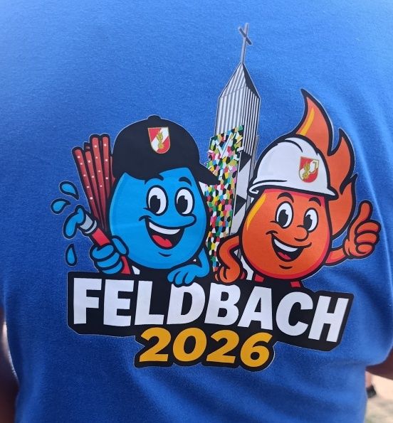 A blue shirt displays a graphic with two cartoon characters and a tall building. The characters are holding tools and smiling. The words 'FELDBACH 2026' are printed below.