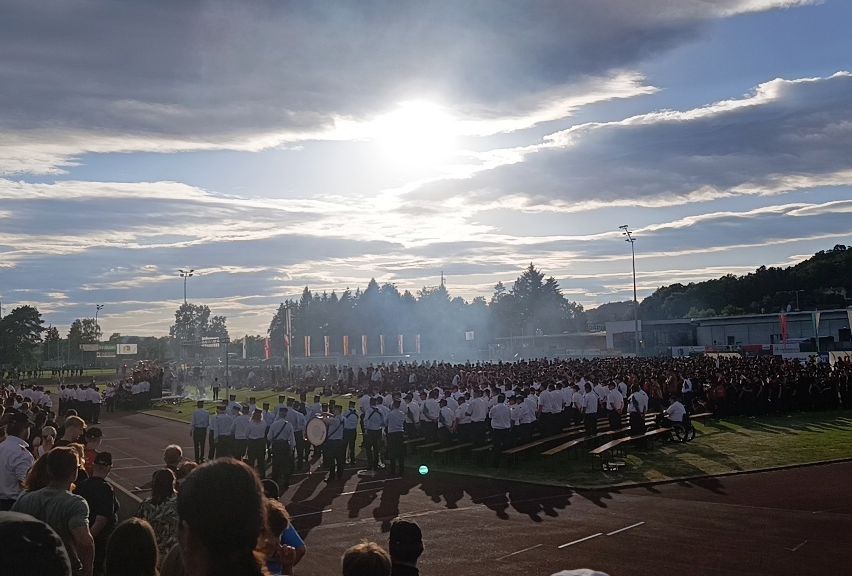 A large crowd gathers on a grassy field under a partly cloudy sky, with a marching band in the middle and spectators on the sides.