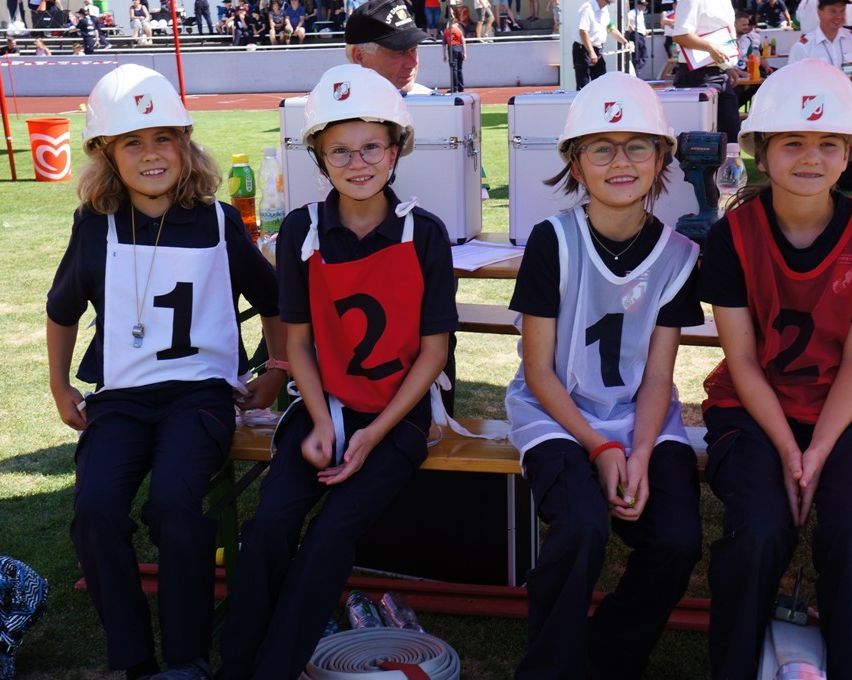 Four children, wearing white hard hats, sit on a bench at a sports event. They are dressed in numbered vests.