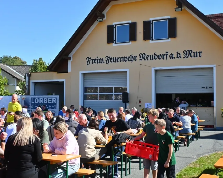 Eine Gruppe von Menschen sitzt an Tischen vor einem Gebäude mit einem Schild, das Freiwillige Feuerwehr Neudorf a.d. Mur steht.