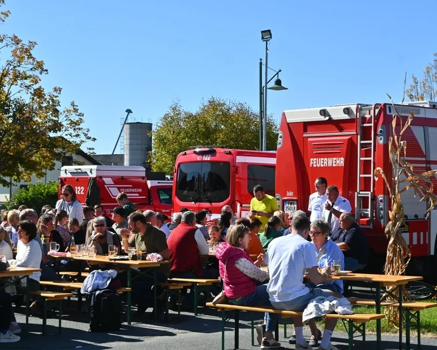 Eine Gruppe von Menschen sitzt an Tischen in einem Außenbereich, möglicherweise einem Park. Es sind rote Feuerwehrfahrzeuge geparkt.