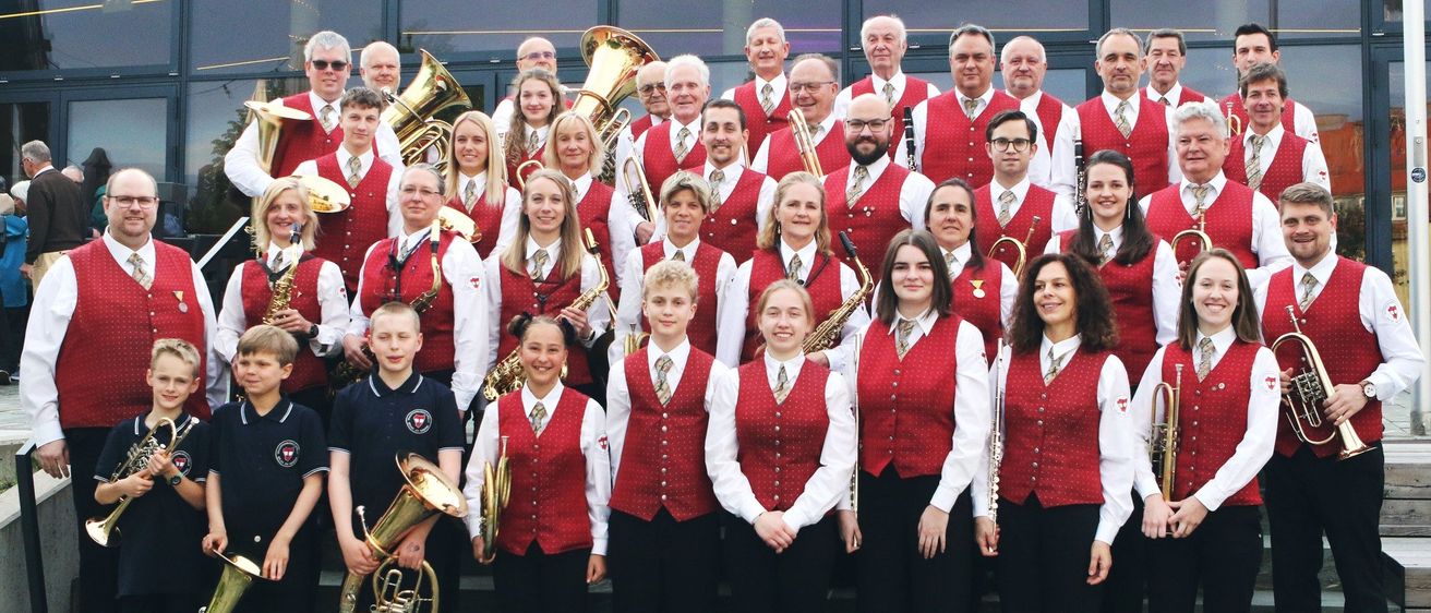 Eine große Gruppe von Menschen in roten Westen und schwarzen Hosen posiert für ein Foto vor einem Gebäude. Sie halten Musikinstrumente wie Trompeten, Saxophone und Tubas.